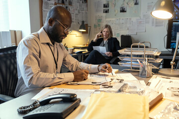 Aufkleber - Serious African American male detective making notes about criminal in his profile while sitting by desk against mature female coworker