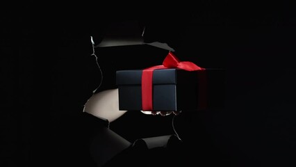 Poster - Black Friday sale. Holiday present. Night shopping. Female hand giving away gift box with red ribbon in breakthrough paper hole ripped wall isolated on dark copy space background.