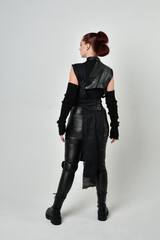 Full length portrait of pretty red haired female model wearing black futuristic scifi leather costume. Dynamic standing poses with gestural hands, facing backwards away from  on a white studio backgro