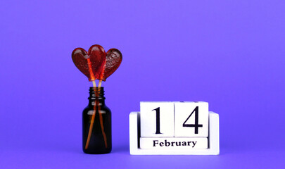 Two red heart shaped lollipops in a small glass bottle and a white wooden calendar with the date February 14 on a purple background. Valentine's Day