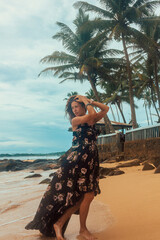 Wall Mural - vacation leisure. young brunette girl in black stylish dress is standing in profile with hands near hair and looking away on a beach coast background near ocean. travel lifestyle concept, free space