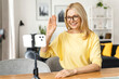© Vadim Pastuh - Senior gray-haired female holding mobile phone and waving hand. Happy smiling mature woman sitting in the kitchen, relaxing and talking to her friend or family via video connection