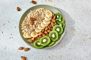 Wall Mural - Healthy Breakfast of tasty granola with kiwi, banana and almond. Slices of exotic fruits and nuts with oat flakes in green plate on the table. Top view.