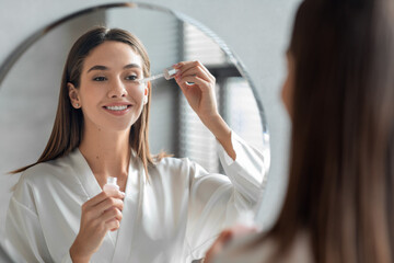 Skin Care Concept. Beautiful Millennial Woman Applying Face Serum Near Mirror