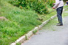A Utility Worker Mows Tall Green Grass With A Petrol Trimmer Mower.