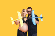 Bearded Man Kisses A Pregnant Girl On A Yellow Background. Couple Playing With Airplanes, Peaceful Sky Concept, Protection From Air Attack