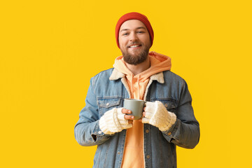 Wall Mural - Young bearded man in warm gloves with cup of hot beverage on yellow background