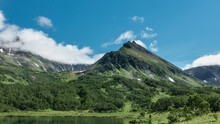 The Picturesque Mountain Range Is Covered With Green Vegetation. Patches Of Melted Snow Are Visible On The Slopes. There Is An Emerald Lake In The Valley. Clouds In The Blue Sky. Kamchatka.