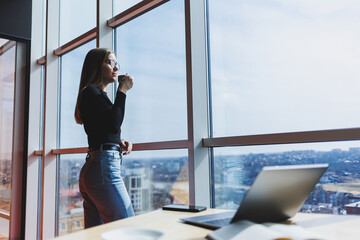 Wall Mural - European woman drinks coffee and looks out the window in a cafe. A young smiling girl in glasses stands near the window. Recreation, leisure and free time. Modern female lifestyle