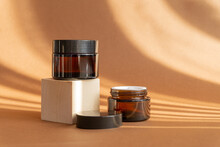 Two Jars Of Amber Glass For Cosmetics On Brown Background In Rays Of Sunlight. Mock-ups Of Containers With Moisturizer For Face And Hands On 3D Geometric Podium. Concept Of Body Care