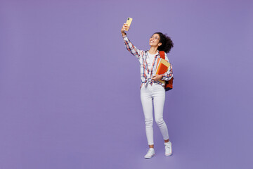 Wall Mural - Full body fun young girl woman of African American ethnicity teen student in shirt doing selfie shot on mobile cell phone isolated on plain purple background. Education in university college concept.