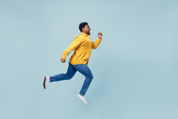 Wall Mural - Full body side view young man of African American ethnicity 20s wear yellow shirt jump high run fast hurry up isolated on plain pastel light blue background studio portrait. People lifestyle concept.