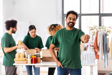 charity, donation and volunteering concept - happy smiling male volunteer pointing to camera and international group of people packing food in boxes at distribution or refugee assistance center