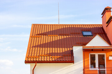 Facade of new white apartment building with red terra cotta tile roof against blue sky. Newly built modern comfortable residential house balcony. Real estate investment. Chimney, small window in roof.