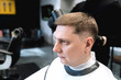 © velirina - Visit to barbershop. Stylish man makes fashionable haircut. Barber,hairdresser,stylist with scissors, comb cutting,dryer.Combing hair.Client,customer sits in dark men's beauty salon covered with cape