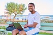 Young smiling male freelancer relaxing on beach, sitting on bench using laptop