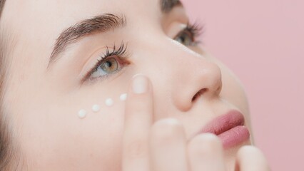 Extreme close-up of young beautiful fit Caucasian skin care woman applies cream under her eye on pink background | Eye cream commercial concept