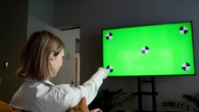 A girl is sitting at home in the living room and watching TV with a green screen Chroma Key while relaxing on the sofa. Changes TV channels with the remote control.