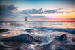 Ocean tide and rocks with lighthouse in the distance seen during colorful sunrise