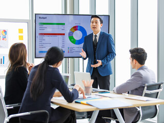 Asian professional successful businessman in formal suit standing showing presenting explaining report investment graph chart data from computer monitor in meeting room to businesswoman colleagues