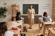© Mediaphotos - Full length portrait of smiling female teacher standing by blackboard in class and talking to group of children, copy space