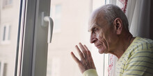Sad Senior Pensioner Looking Through Window And Touching Glass With Hand