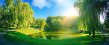 Panoramic Natural Spring Landscape. A Small Lake In Green Park With Trees Against Blue Sky With Bright Sun.