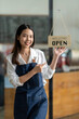 © crizzystudio - Happy waitress stands at the entrance of the restaurant showing off a sign of the store open. persuade customers to enter the store The smiling owner in a blue apron stood at the entrance to the cafe