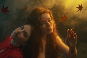 Art studio portrait made through the wet glass with red autumn leaves. Two beautiful redhead freckled women with long natural curly hair posing in smoke, darkness and warm light. Film grain effect
