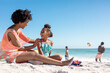 Cheerful african american mother and daughter sitting together while father and son playing at beach