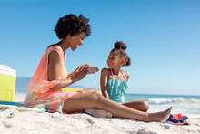 Happy African American Woman Applying Suntan Lotion On Daughter While Sitting At Beach On Sunny Day