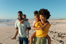 Happy African American Parents Giving Piggyback To Children While Walking At Beach On Sunny Day