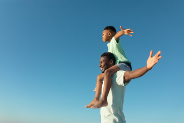 happy african american boy on father's shoulders by copy space against clear blue sky