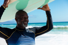 Smiling African American Bald Retired Senior Man Carrying Surfboard On Head At Beach