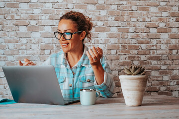 Wall Mural - Hipter young woman talking in video call conference onine on laptop. People at work with computer. Bricks background and happy lady speaking and enjoying communication modern job