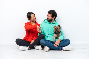 Wall Mural - Young caucasian couple sitting on the floor with their pet isolated on white background handshaking after good deal