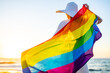 © yurakrasil - woman with straw white hat holding a gay pride rainbow flag blowing in the wind on beach with golden sunset