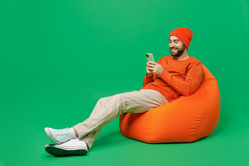 Full body young fun happy caucasian man 20s wear orange sweatshirt hat sit in bag chair hold in hand use mobile cell phone isolated on plain green background studio portrait. People lifestyle concept.