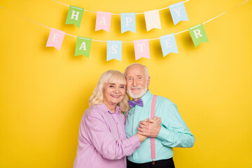 Poster - Portrait of two attractive cheerful grey-haired spouses hugging easter day isolated over bright yellow color background