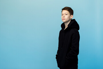 A thoughtful boy in a black hoodie and jeans looks at the camera on a monochrome blue background. There is a place for the inscription.