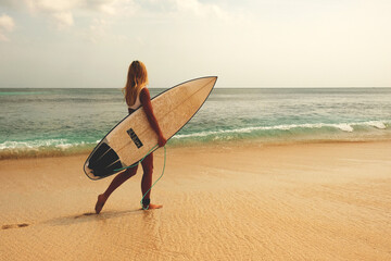 blonde surfing girl walking along a sandy beach with a dock for surfing on the background of a tropical wave