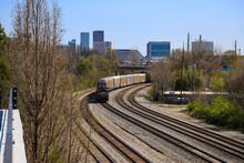 A Train Stopped On The Railroad Tracks Surrounded By Gravel, Lush Green Trees And Plants With Skyscrapers And Office Buildings In The Cityscape With Blue Sky In Atlanta Georgia USA