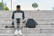 © Oscar - front view of an african american man sitting on a staircase working on his laptop. hipster man listening to music with yellow headphones.
