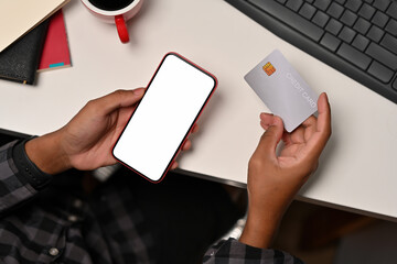 Top view with man's hand holding a smartphone with blank white screen and holding a credit card while sitting at the office desk, for finance, business and technology concept.