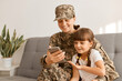 © sementsova321 - Indoor shot of smiling happy delighted family sitting on sofa and using phone, woman wearing camouflage uniform and cap posing with her daughter looking at mobile phone.