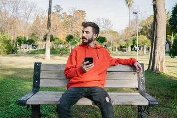 Wall Mural - Young man sitting on a park bench and looking away with phone. Holding smartphone, thinking over text message for online chat