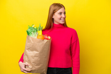 Wall Mural - Young redhead woman holding a grocery shopping bag isolated on yellow background looking to the side and smiling