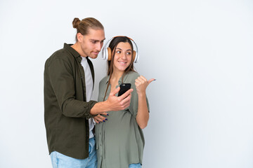 Wall Mural - Young caucasian couple isolated on white background pregnant and listening music while pointing side