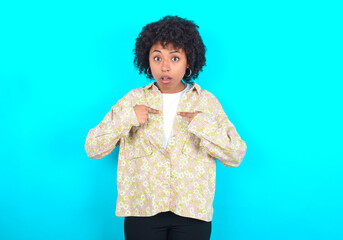 Wall Mural - Embarrassed young girl with afro hairstyle wearing floral shirt indicates at herself with puzzled expression, being shocked to be chosen to participate in competition, hesitates about something