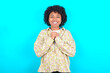 © Roquillo - Positive young girl with afro hairstyle wearing floral shirt over blue background smiles happily, glad to receive pleasant news from interlocutor, keeps hands together. People emotions concept.